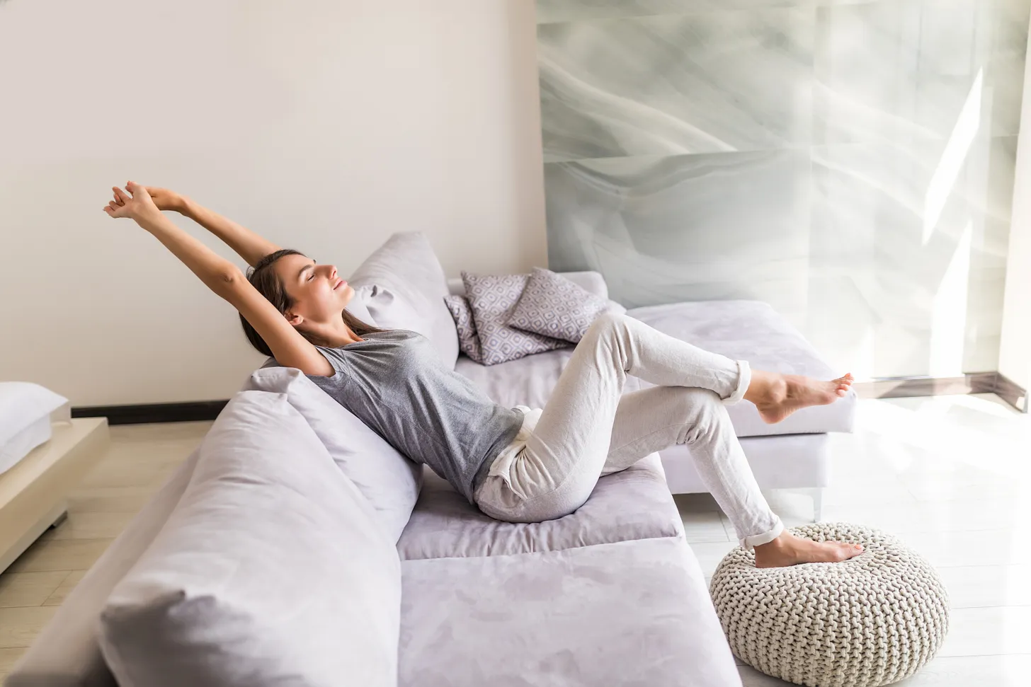 mulher jovem a relaxar e a alongar os braços, sentada num sofá dentro de casa.