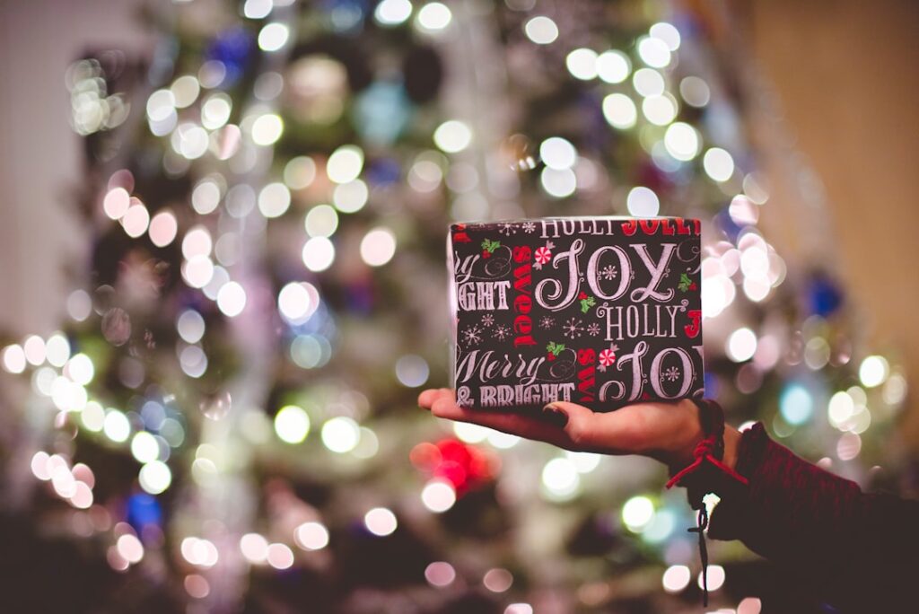 mão feminina a segurar um presente embrulhado com árvore de natal iluminada no fundo